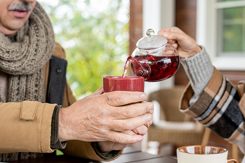 Hand of senior woman with teapot pouring herbal tea into ceramic mug held by her husband over table during breakfast by country house