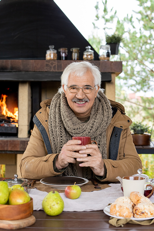 Cheerful senior man in warm jacket and knitted scarf holding mug with hot tea while sitting by served table in front of camera