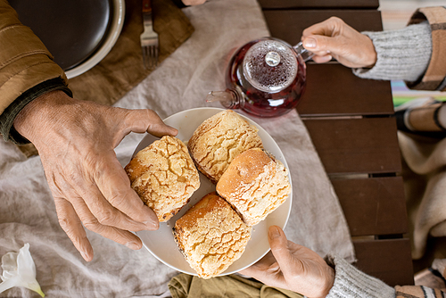 Hand of aged man taking fresh homemade bun from plate held by his careful wife over served table while both having breakfast