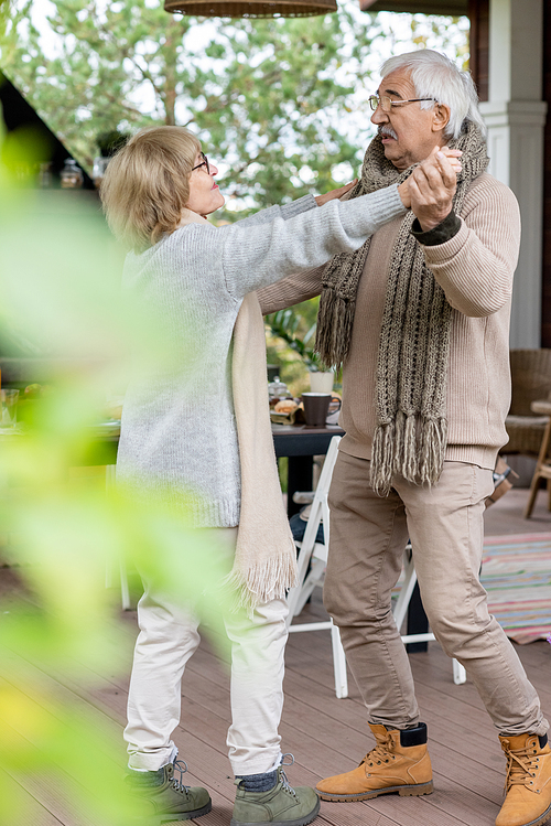 Affectionate senior couple in warm stylish casualwear dancing on wooden floor of patio by their country house against served table