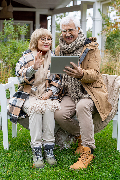 Elderly husband and wife with digital tablet communicating in video chat while sitting on bench on green lawn in front of their country house