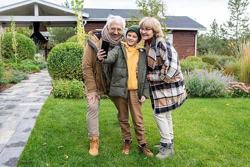 Happy youngster in casualwear and his affectionate grandparents looking in smartphone camera with smiles while making selfie