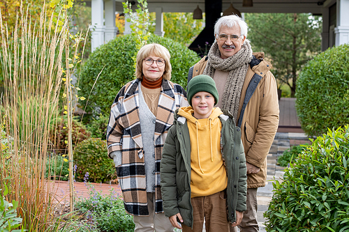 Happy youngster in casualwear and his affectionate grandparents looking at you with smiles while standing in front of camera in the garden