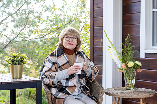 Retired blond female in eyeglasses and warm stylish checkered coat holding mug with hot tea while sitting in armchair by country house