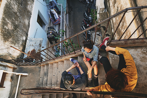 High angle view of stylish Asian teenagers hanging out in urban slums: they sitting on stairs of shabby apartment house and talking to each other