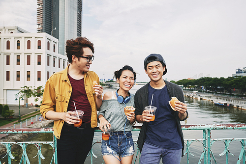 Happy young people with wide smiles holding sandwiches and plastic cups of iced tea in hands while leaning against bridge railing