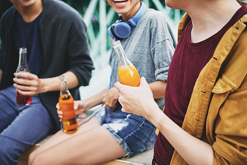 Group of cheerful friends chatting animatedly with each other while sitting on bridge railing and drinking delicious cocktails, close-up shot