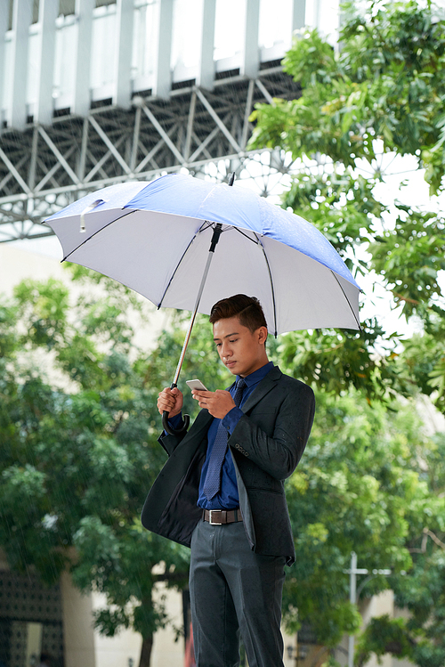 Portrait of handsome young businessman using smartphone outdoors standing in street under umbrella