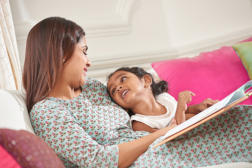 Happy Indian mother and daughter lying in bed and reading together