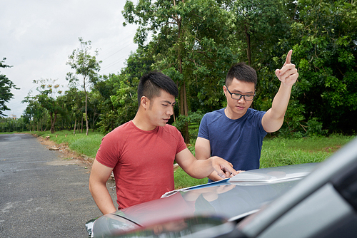 Two handsome Vietnamese friends parked car on side of road and reading paper map with concentration