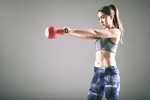 Pretty sportswoman doing exercises with heavy kettlebell, isolated on grey