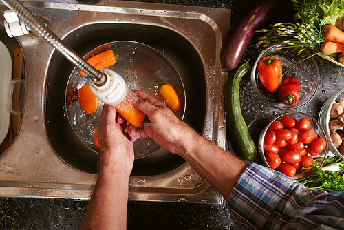 Hands of man washing orange bell peppers in kitchen sink