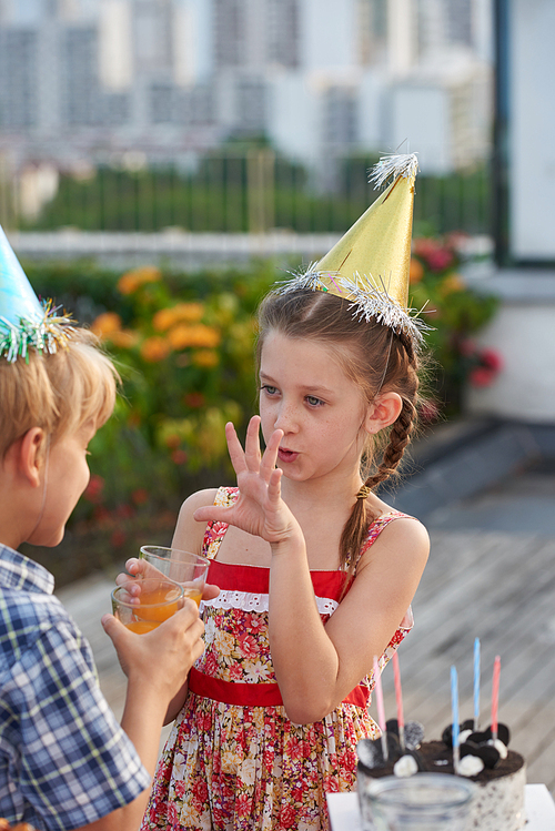 Pretty dark-haired little girl gesticulating and talking to her best friend while having fun at outdoor birthday party