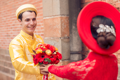 Portrait of cheerful Caucasian groom wearing traditional Vietnamese costume looking at his bride with toothy smile while passing her colorful wedding bouquet, facade of building on background