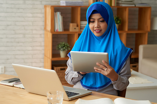Concentrated young woman in hijab sitting at table with laptop and using digital tablet