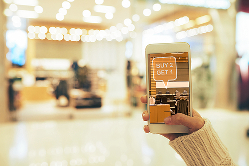 Close-up shot of young woman using smartphone with augmented reality app while doing shopping at clothing shop, blurred background