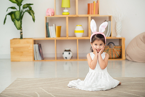 Full length portrait of adorable little girl wearing white dress and bunny ears sitting on cozy carpet and posing for photography, she holding hands on cheeks, interior of living room on background