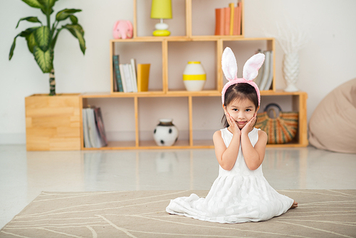 Full length portrait of adorable little girl wearing white dress and bunny ears sitting on cozy carpet and posing for photography, she holding hands on cheeks, interior of living room on background