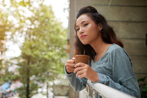 Beautiful young Vietnamese woman with cup of tea looking out of her balcony