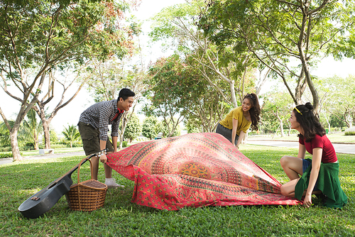 Friends putting blanket on grass to have picnic