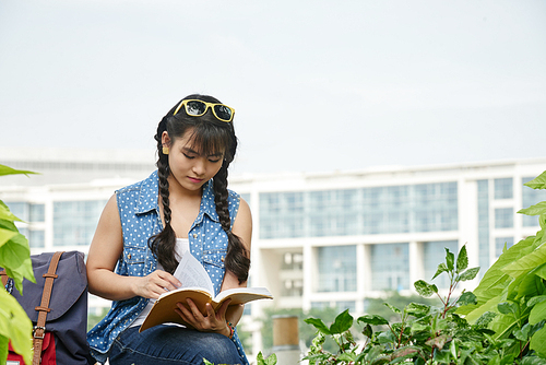 Pretty young student sitting on bench in university campus and preparing for exam, multi-storey building on background