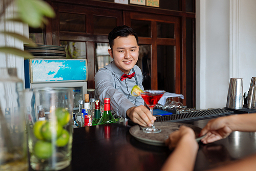 Handsome young barman with toothy smile putting cocktail glass on tray while standing at bar counter, portrait shot
