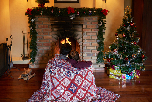 Rear view of loving couple wrapped in blanket sitting in front of fireplace on Christmas Eve