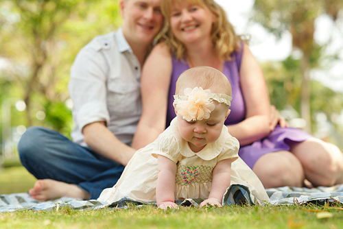 Portrait of cute toddler wearing dress and headband crawling on green lawn while her middle-aged parents sitting on checked plaid behind her and posing for photography, sunny park on background