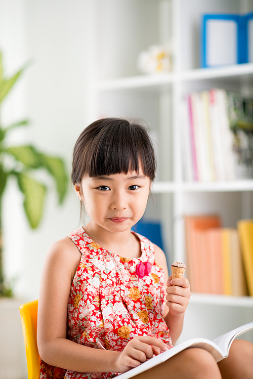 Adorable Vietnamese girl posing for photography with warm smile while eating appetizing ice-cream and reading fairy-tales, interior of cozy bedroom on background