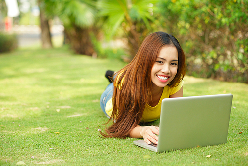 Cheerful Asian student wearing T-shirt and jeans lying on green lawn at public park while doing homework with help of laptop, portrait shot