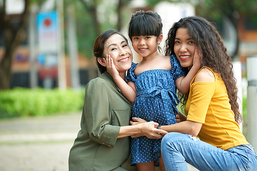 Family portrait of attractive middle-aged woman and her mature mother sitting on haunches while embracing cute little girl and  with wide smiles, green public park on background