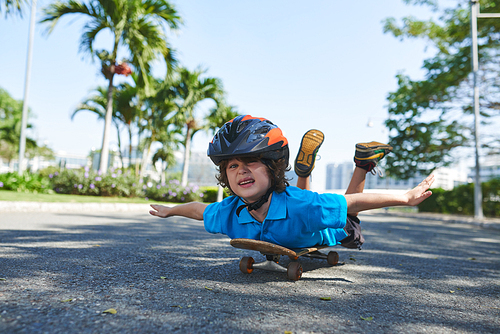 Curly little boy in blue polo T-shirt lying on skateboard on his belly and pretending to fly in sunny park
