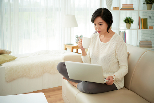 Portrait of Asian woman relaxing at home sitting on comfortable sofa holding laptop and drinking cocoa
