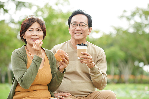 Happy senior Asian couple eating buns and drinking take-out coffee in the park