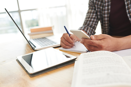 Hands of student reading something on smartphone when taking notes