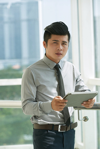 Pensive Asian businessman with stylish haircut standing against panoramic window and working on his project with help of digital tablet