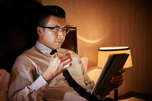 Asian businessman lying in bed and reading something on digital tablet at night