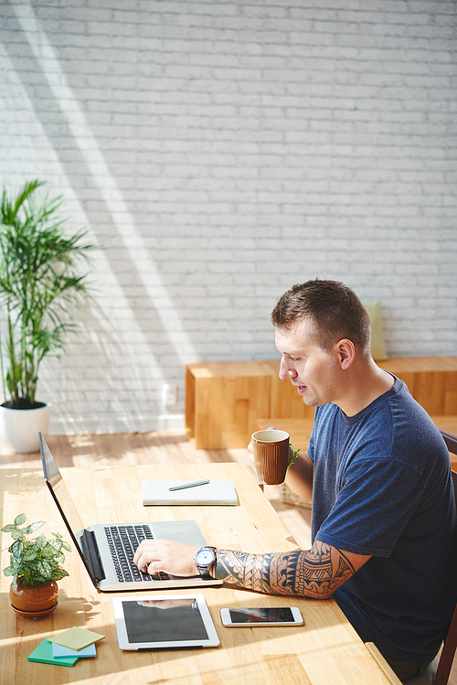 Young man with tattoo on his arm drinking coffee and working on laptop