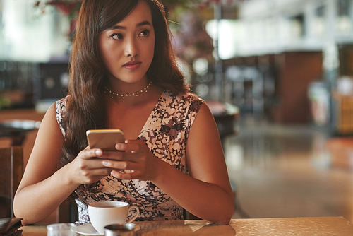 Attractive young Chienese woman having cup of coffee and checking her smartphone