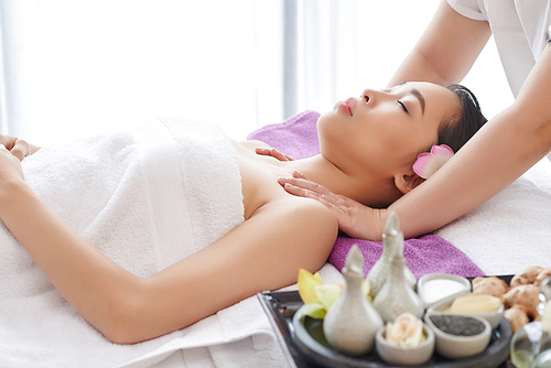 Beautiful Asian woman lying on massage table with closed eyes and enjoying procedure, waist-up portrait