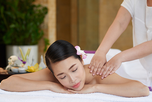 Unrecognizable massage therapist carrying out procedure in modern beauty salon, pretty young woman relaxing on massage table with closed eyes