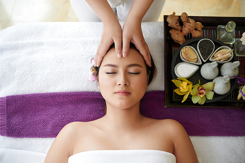 Spending weekend in massage salon: pretty young woman lying on table with closed eyes while therapist massaging her head