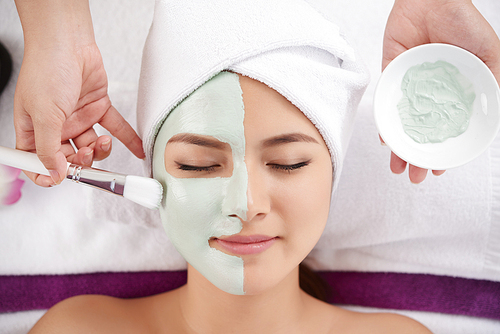 Beautician applying clay face mask on face of pretty woman