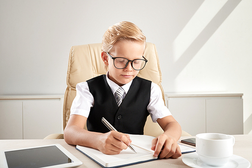 Serious little business executive in glasses writing ideas into textbok