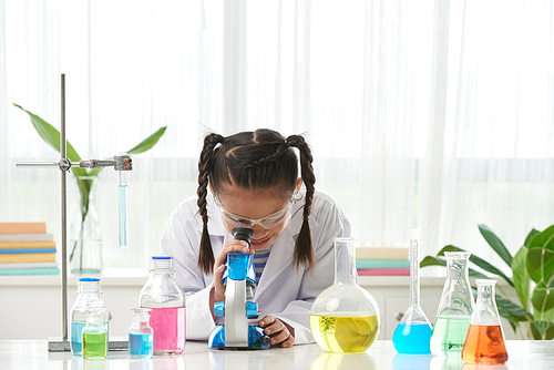 Asian schoolgirl in goggles and white coat looking into microscope