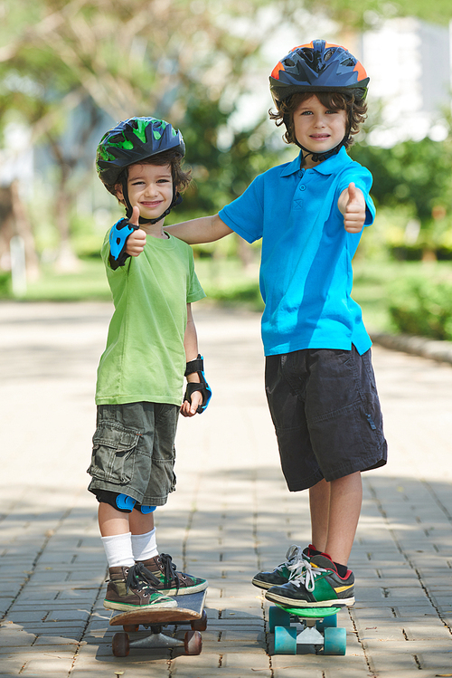 Full-length portrait of handsome little boys with thumbs up standing on skateboards and  in sunny green park