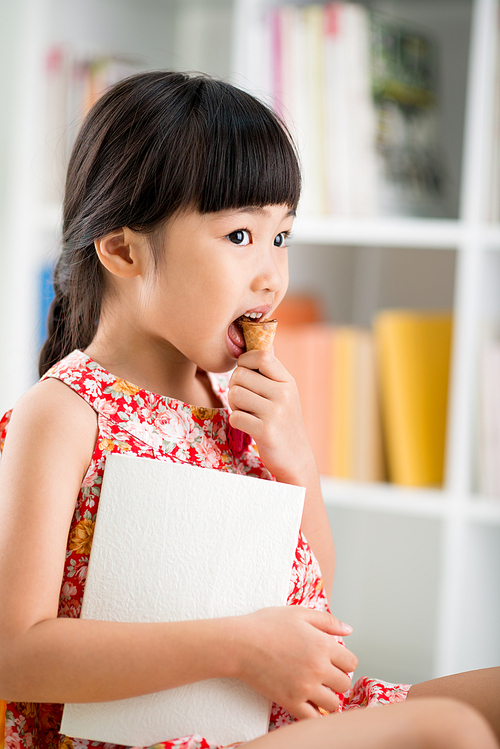 Profile view of adorable little girl wearing colorful dress looking away while eating appetizing ice cream cone, blurred background