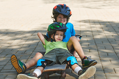 Two cute preschoolers in colorful helmets and t-shirts sitting on skateboard one after another and  cheerfully
