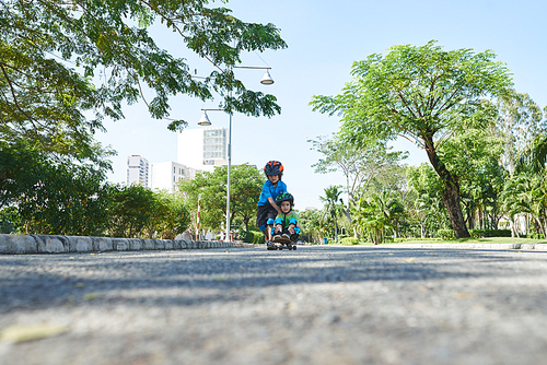 Cute little boy in helmet and knee pads riding skateboard while sitting on it, his elder brother pushing him down the road, surface level shot