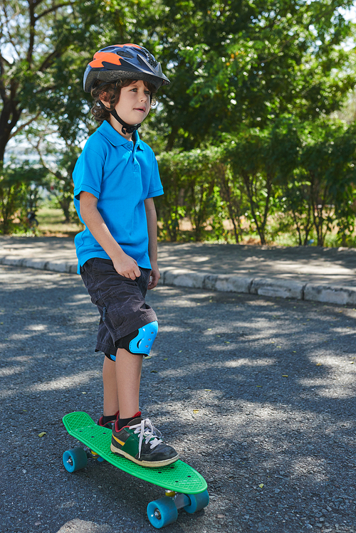 Thoughtful curly little boy standing with one leg on bright green penny board and looking into distance in summer park